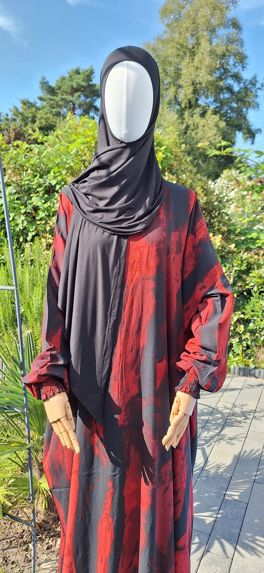 Abaya Qurtuba in kräftigem Rot-Schwarz-Muster, aufgenommen vor grüner Naturkulisse.