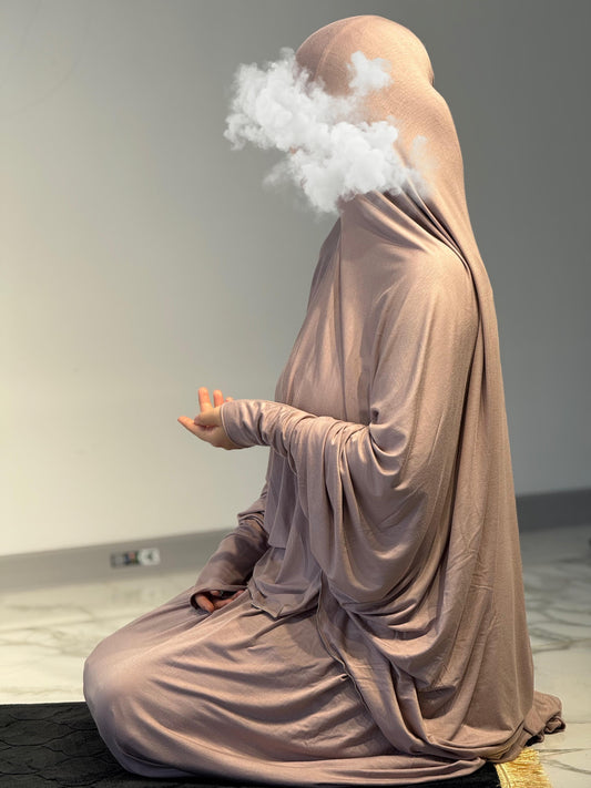 Frau im Jilbab Beige Sahra in sitzender Gebetshaltung auf schwarzer Gebetsmatte, weicher Stoff fällt fließend bis zum Boden.