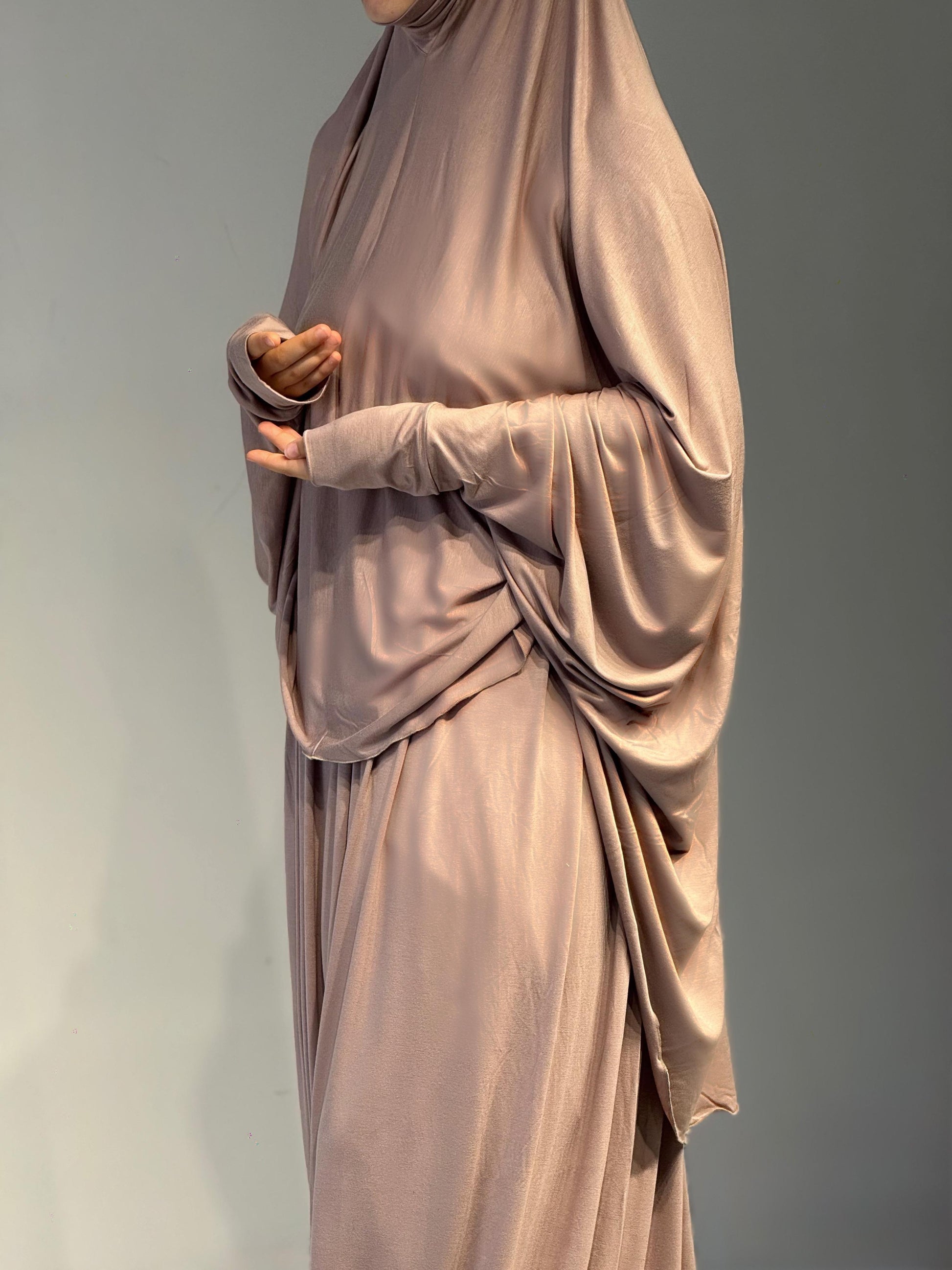 Frau steht seitlich im Jilbab Beige Sahra, mit einem fließenden Fall entlang der Silhouette. Leichte Drapierungen sind sichtbar.