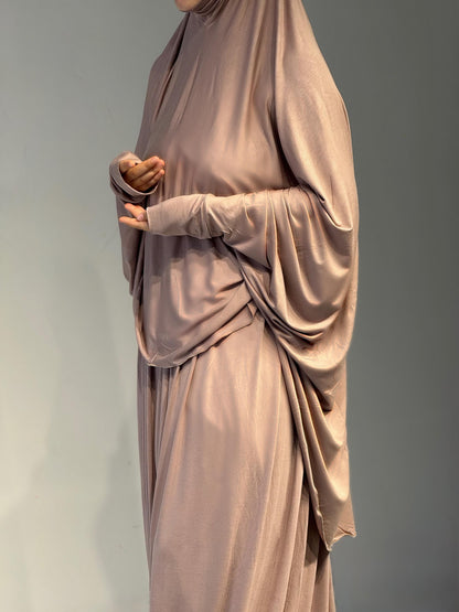 Frau steht seitlich im Jilbab Beige Sahra, mit einem fließenden Fall entlang der Silhouette. Leichte Drapierungen sind sichtbar.