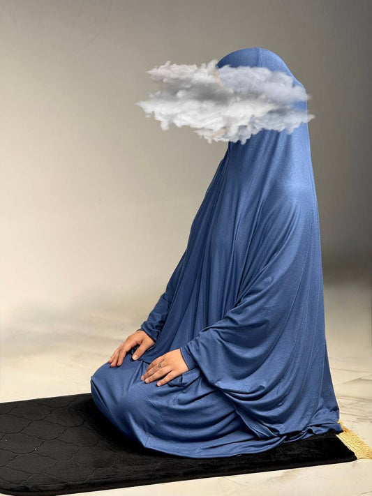 Frau im Jilbab Azraq Ocean im Gebet sitzend auf schwarzer Gebetsmatte, edler blauer Stoff fällt weich bis zum Boden.